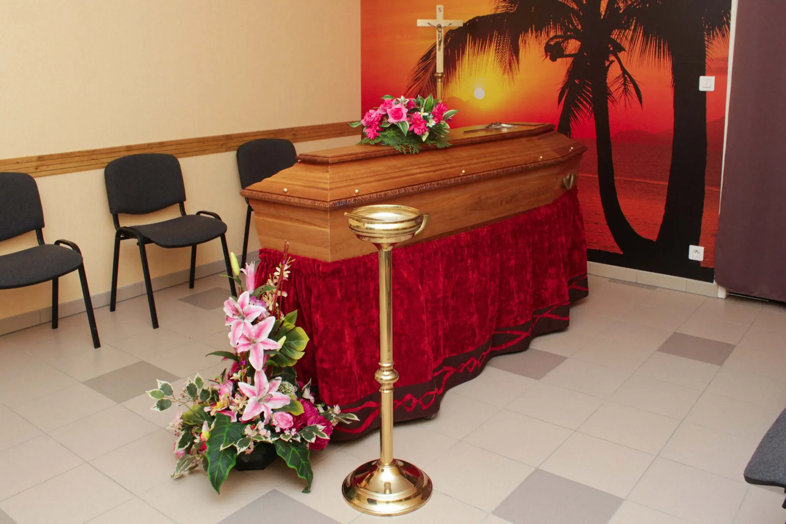 Cercueil en bois fleuri dans une chambre funéraire aménagée avec chaises et crucifix.